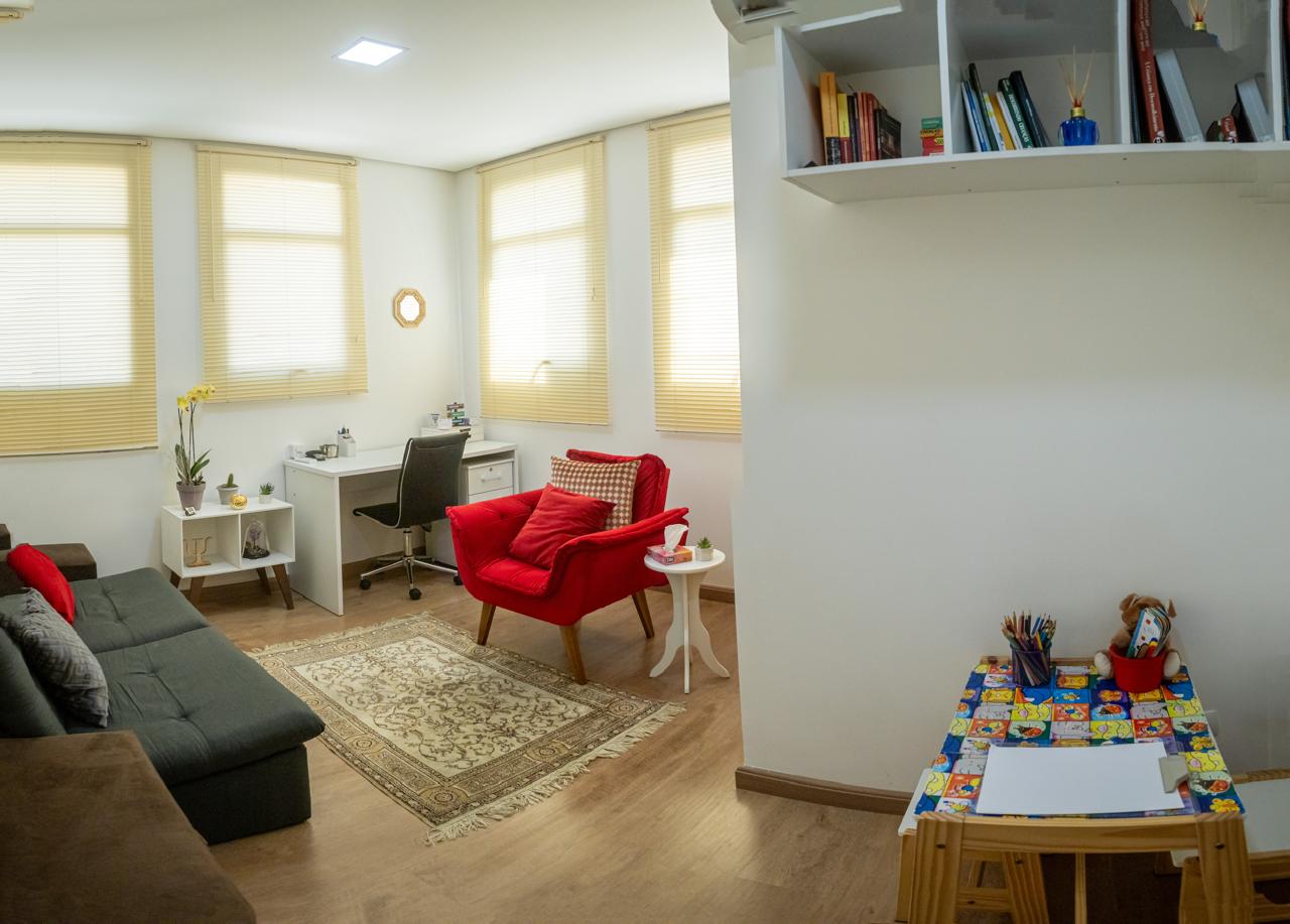 Sala de atendimento da Clínica JF com poltrona vermelha, sofá cinza, mesa de trabalho e estante de livros